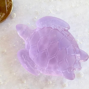 Puede incluir: Una pequeña figura de tortuga marina de color lavanda con un caparazón texturizado. Una moneda de diez centavos estadounidense está al lado de la tortuga para comparar el tamaño. La moneda está fechada en 1999. La tortuga está hecha de un material translúcido.