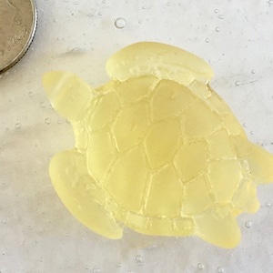 Puede incluir: Una pequeña figura de tortuga marina de color amarillo pálido con un caparazón texturizado, junto a una moneda de diez centavos estadounidense para comparar el tamaño. La tortuga tiene aletas y cabeza visibles. El fondo es una superficie clara y moteada.