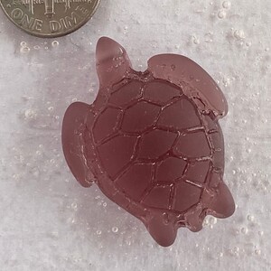Puede incluir: Una pequeña figura de tortuga marina de vidrio púrpura. La tortuga tiene un caparazón detallado con un diseño texturizado.