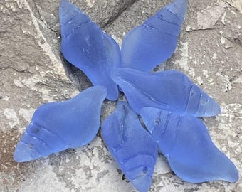 May include: Six blue glass sea shells in various shapes and sizes. The shells are arranged in a starburst pattern on a gray, textured surface.