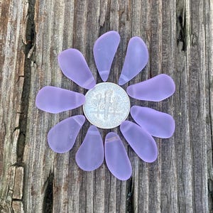 Könnte beinhalten: Lavendelfarbene Seeglasstücke, die in Form einer Blume um einen silbernen Dime angeordnet sind. Das mattierte Seeglas liegt auf einer verwitterten Holzoberfläche. Der Dime hat einen Durchmesser von ca. 1,79 cm.