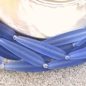 May include: Close-up of a necklace featuring elongated, frosted blue beads. The beads are strung together with small, clear beads. The necklace is set against a neutral background, highlighting the cool blue tones of the jewelry.