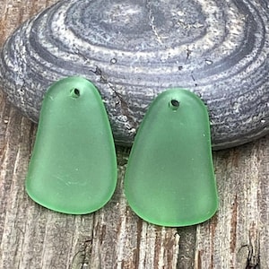 Puede incluir: Dos colgantes de vidrio marino en un suave tono verde, en forma de lágrima con un pequeño agujero en la parte superior para ensartar. Los colgantes descansan sobre una superficie de madera envejecida, con una piedra rayada gris y blanca en el fondo.