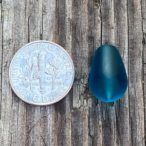 May include: A smooth, matte, teardrop-shaped bead in a deep teal blue color. The bead is positioned next to a United States dime, which has a diameter of 0.705 inches, providing a clear visual reference for the size of the bead.