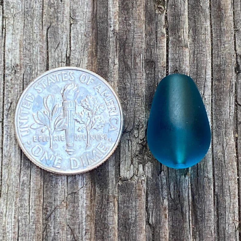 May include: A smooth, matte, teardrop-shaped bead in a deep teal blue color. The bead is positioned next to a United States dime, which has a diameter of 0.705 inches, providing a clear visual reference for the size of the bead.