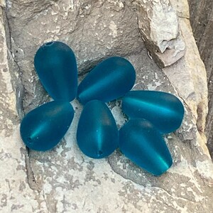 May include: A collection of six teardrop-shaped beads in a matte teal blue finish. Each bead features a smooth, frosted surface texture and a small hole at the narrow end for stringing. The beads are arranged in a cluster on a textured surface.