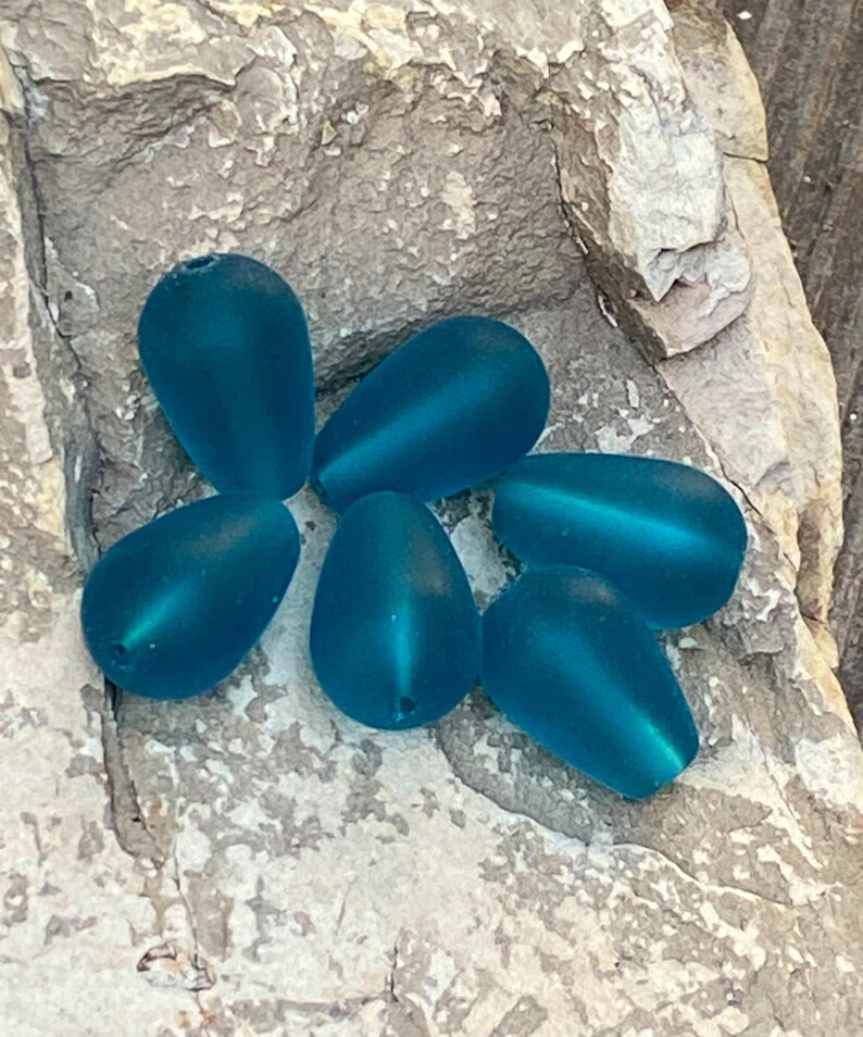 May include: A collection of six teardrop-shaped beads in a matte teal blue finish. Each bead features a smooth, frosted surface texture and a small hole at the narrow end for stringing. The beads are arranged in a cluster on a textured surface.
