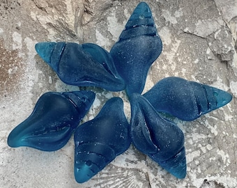 May include: Six blue glass seashells in the shape of a conch shell. The shells are arranged in a circle on a gray, textured surface.