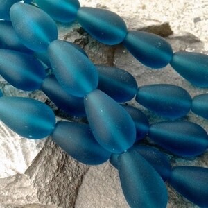 May include: A collection of teardrop-shaped beads in a deep teal blue color. Each bead features a smooth, frosted matte finish, creating a soft texture. The beads are arranged in a cluster, showcasing their uniform shape and consistent blue hue.