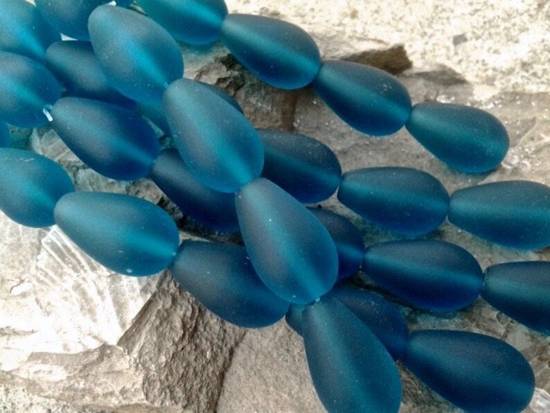 May include: A collection of teardrop-shaped beads in a deep teal blue color. Each bead features a smooth, frosted matte finish, creating a soft texture. The beads are arranged in a cluster, showcasing their uniform shape and consistent blue hue.