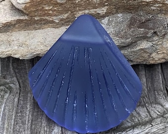 May include: A deep blue glass sea shell with a textured surface. The shell is shaped like a scallop and has a smooth, rounded edge.
