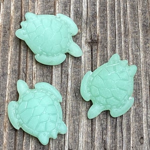 May include: Three light green glass sea turtle figurines on a wooden surface.