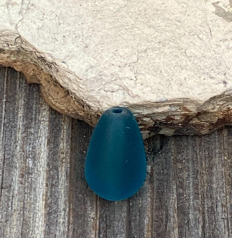 May include: A single, teardrop-shaped bead in a deep, matte teal color. The bead features a smooth, frosted surface texture and a small, centered hole at the top for threading. The object is solid in color and has a rounded, symmetrical base.