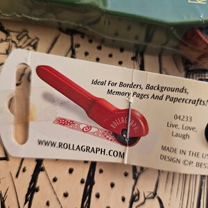 May include: A red and white rolling tool with a white handle and a red wheel. The tool is labeled "Rollagaph" and has the text "Ideal For Borders, Backgrounds, Memory Pages And Papercrafts!" on the packaging. The packaging also includes the text "04233 Live, Love, Laugh Made In The US Design OP. BEST" and the website address "www.rollagaph.com".