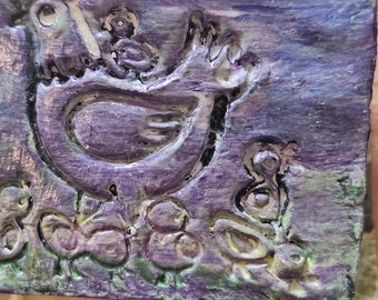 Puede incluir: Un azulejo de cerámica morado con un diseño en relieve de una gallina y pollitos. El azulejo tiene una superficie texturizada y un esmalte verde claro y amarillo.