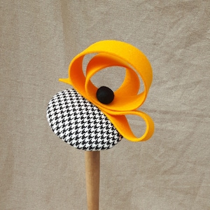 May include: A black and white houndstooth hat with a bright yellow felt accent. The hat is displayed on a wooden stand.