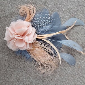 Peut inclure: Une fleur en tissu rose p&acirc;le avec un embellissement en plumes et filet gris et beige.