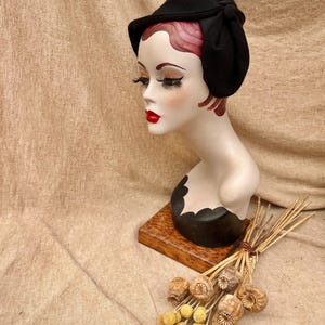 K&ouml;nnte beinhalten: Ein schwarzer Filzhut mit Schleife an der Seite. Der Hut sitzt auf einem Mannequin-Kopf mit roten Haaren. Das Mannequin steht auf einem Holzsockel mit getrockneten Blumen.