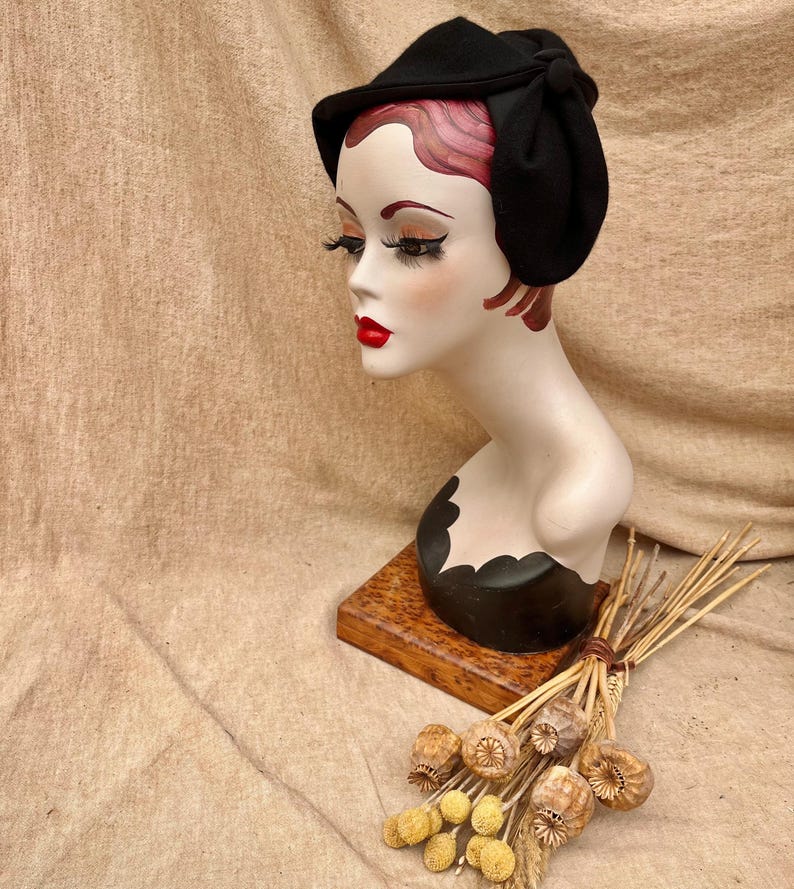 K&ouml;nnte beinhalten: Ein schwarzer Filzhut mit Schleife an der Seite. Der Hut sitzt auf einem Mannequin-Kopf mit roten Haaren. Das Mannequin steht auf einem Holzsockel mit getrockneten Blumen.