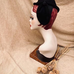 K&ouml;nnte beinhalten: Ein schwarzer Filzhut mit einer gro&szlig;en Schleife und einem kleinen Knopf an der Seite. Der Hut sitzt auf einem Mannequin-Kopf.