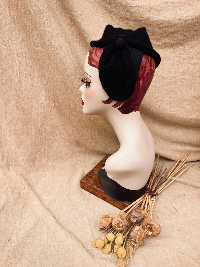 K&ouml;nnte beinhalten: Ein schwarzer Filzhut mit einer gro&szlig;en Schleife und einem kleinen Knopf an der Seite. Der Hut sitzt auf einem Mannequin-Kopf.