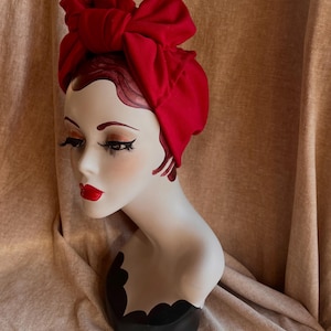 Op de afbeelding: Een rode turban met een grote strik bovenop. De turban is gemaakt van een zachte, rekbare stof en is perfect om elke outfit een vleugje stijl te geven.