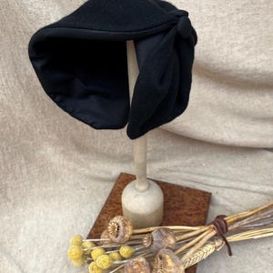 K&ouml;nnte beinhalten: Ein schwarzer Filzhut im Cloche-Stil mit einer gro&szlig;en Schleife und einem Knopf als Akzent. Der Hut wird auf einem Holzst&auml;nder pr&auml;sentiert.