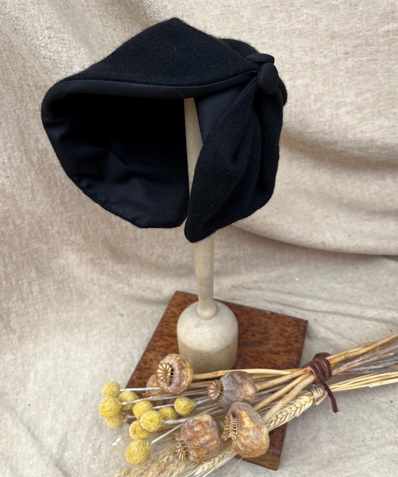 K&ouml;nnte beinhalten: Ein schwarzer Filzhut im Cloche-Stil mit einer gro&szlig;en Schleife und einem Knopf als Akzent. Der Hut wird auf einem Holzst&auml;nder pr&auml;sentiert.