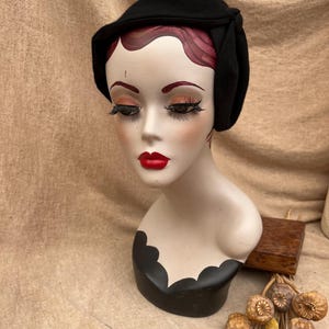 K&ouml;nnte beinhalten: Ein schwarzer Filzhut im Cloche-Stil mit einer gerafften Detail auf der Seite. Der Hut sitzt auf einem Mannequin-Kopf mit roten Lippen und roten Haaren.