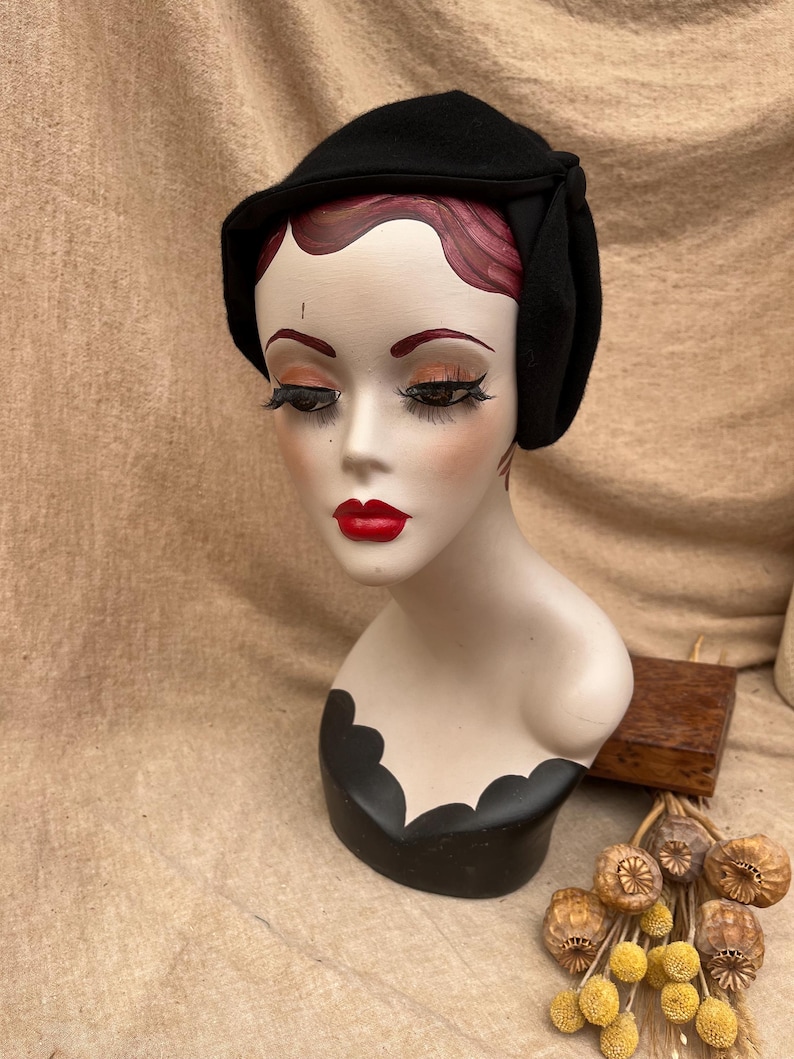 K&ouml;nnte beinhalten: Ein schwarzer Filzhut im Cloche-Stil mit einer gerafften Detail auf der Seite. Der Hut sitzt auf einem Mannequin-Kopf mit roten Lippen und roten Haaren.
