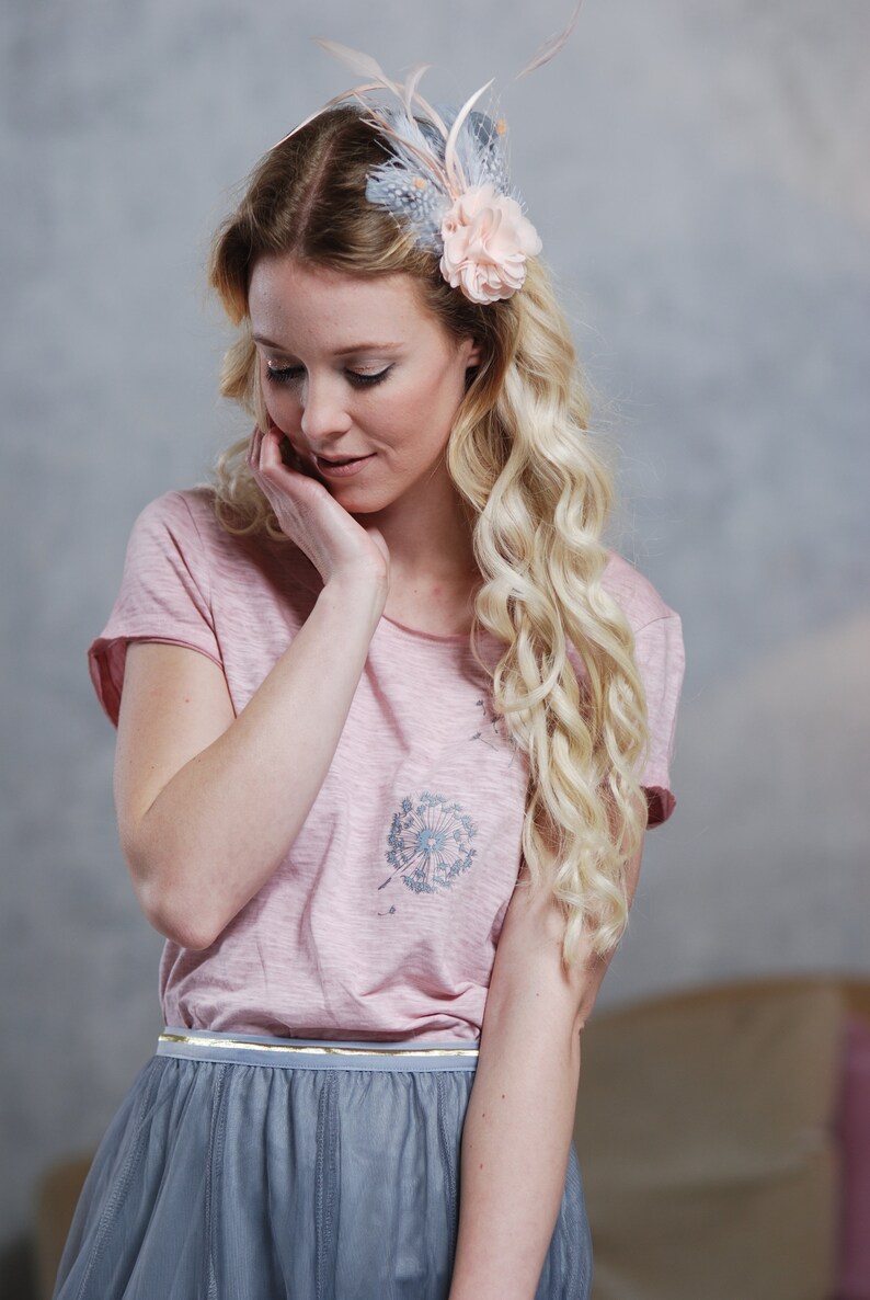 Peut inclure: Une femme portant un t-shirt rose avec un motif de pissenlit et une jupe en tulle grise avec une ceinture dor&eacute;e. Elle porte une barrette de cheveux en fleur rose clair avec des plumes.