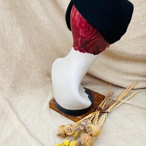 K&ouml;nnte beinhalten: Ein schwarzer Filzhut im Turbanstil auf einem wei&szlig;en Mannequin-Kopf. Das Mannequin steht auf einem Holzsockel. Im Vordergrund befinden sich getrocknete Blumen.