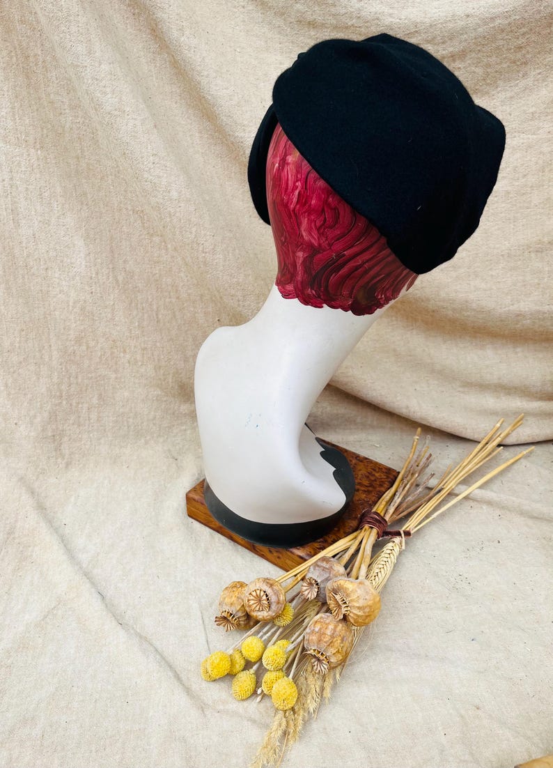 K&ouml;nnte beinhalten: Ein schwarzer Filzhut im Turbanstil auf einem wei&szlig;en Mannequin-Kopf. Das Mannequin steht auf einem Holzsockel. Im Vordergrund befinden sich getrocknete Blumen.