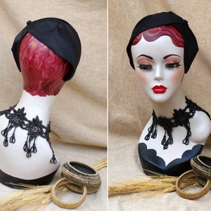 May include: A black satin hat with a bow on the side, displayed on a mannequin with a black beaded necklace. The mannequin is wearing a red wig and is positioned against a beige background.