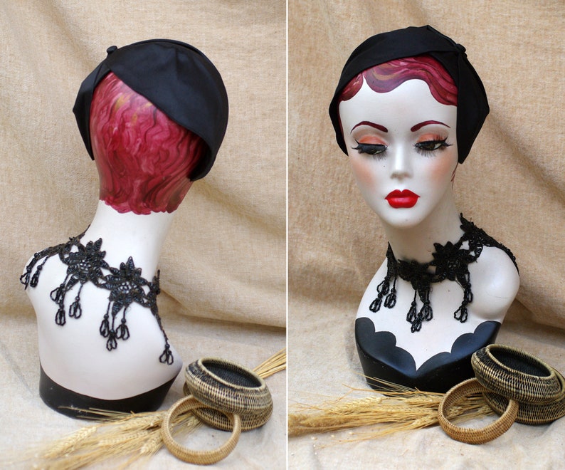 May include: A black satin hat with a bow on the side, displayed on a mannequin with a black beaded necklace. The mannequin is wearing a red wig and is positioned against a beige background.