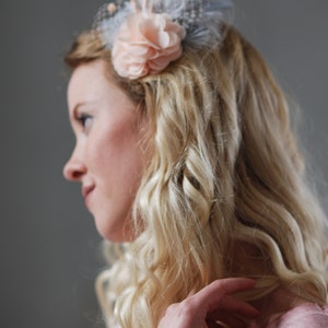 Peut inclure: Une femme avec de longs cheveux blonds ondul&eacute;s portant une barrette de cheveux en forme de fleur rose p&acirc;le avec des plumes bleues et une base en maille bleu clair.