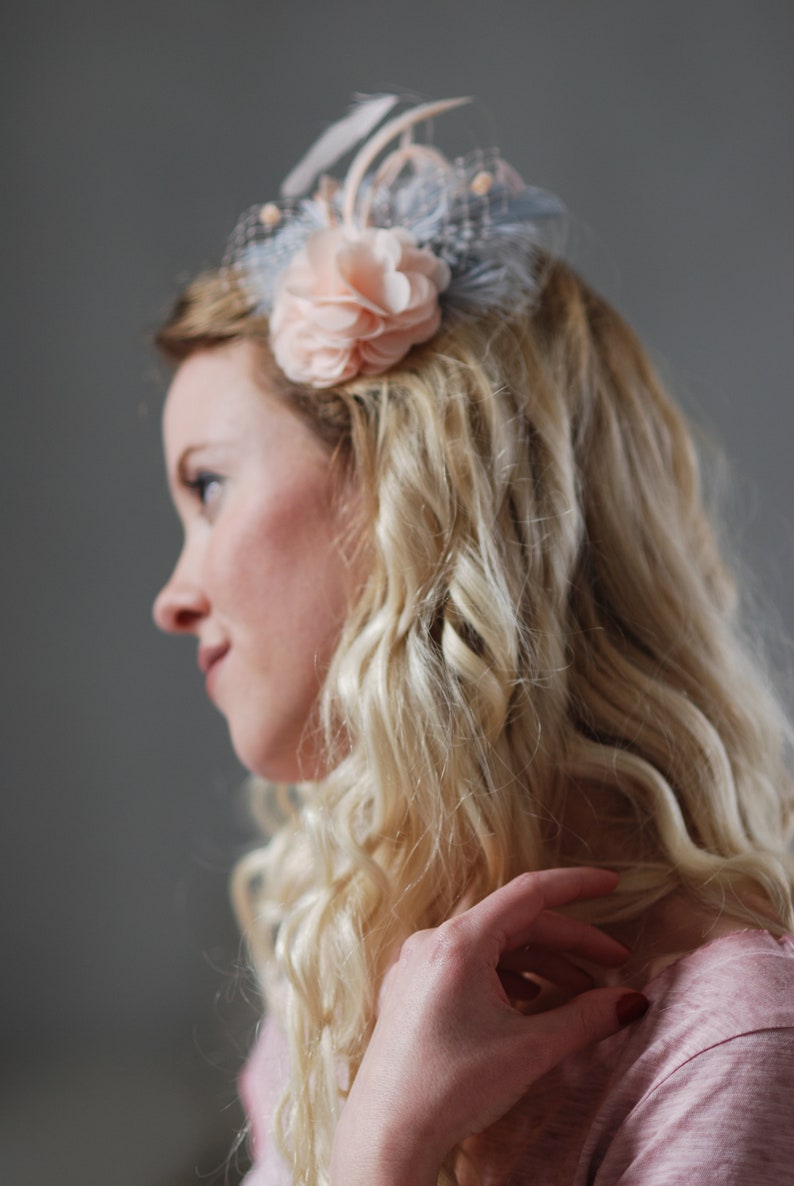 Peut inclure: Une femme avec de longs cheveux blonds ondul&eacute;s portant une barrette de cheveux en forme de fleur rose p&acirc;le avec des plumes bleues et une base en maille bleu clair.