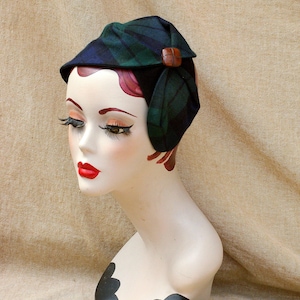 Könnte beinhalten: Ein Hut aus grünem und schwarzem kariertem Stoff mit einem braunen Knopf als Akzent. Der Hut wird auf einem Mannequin-Kopf getragen.