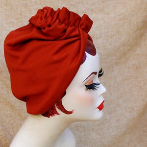 Könnte beinhalten: Ein roter Turban aus weichem Stoff mit einem gerafften Detail an der Oberseite. Der Turban wird von einer Puppe mit bemaltem Gesicht und langen Wimpern getragen.