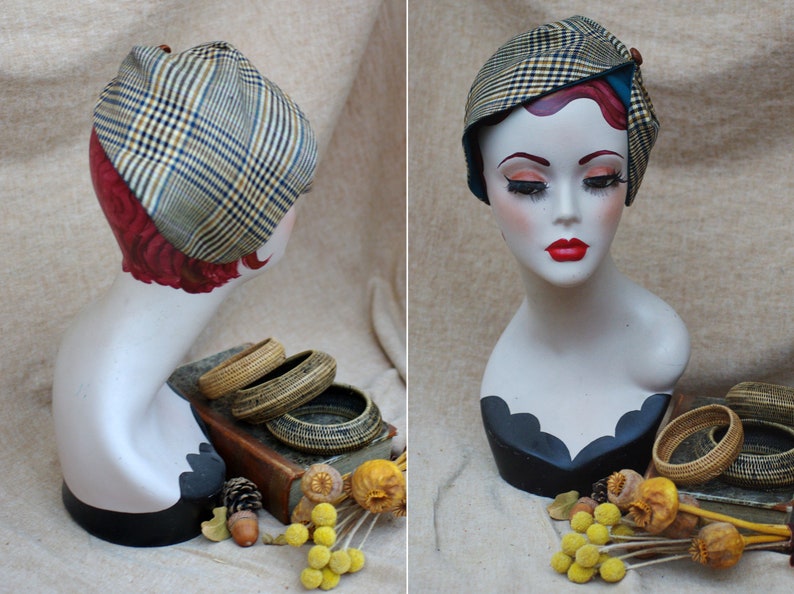 May include: A mannequin head wearing a brown and beige plaid hat with a blue trim. The hat is tilted slightly to the side. The mannequin is wearing a black and white patterned dress.