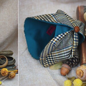May include: A teal and plaid fabric turban with a wooden button. The turban is folded and displayed on a fabric surface with a book, acorns, and dried flowers.