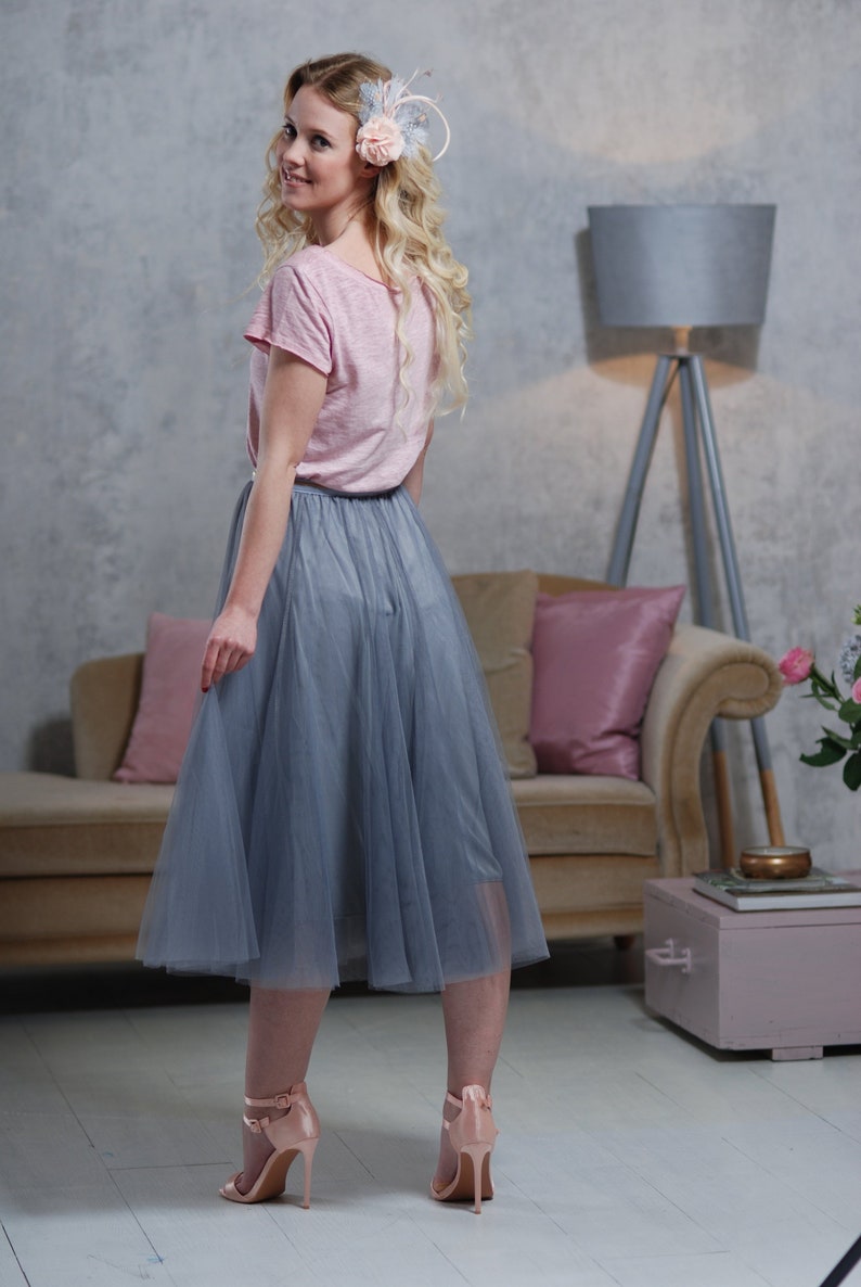 Peut inclure: Une femme portant un t-shirt rose et une jupe en tulle grise avec un clip &agrave; fleurs dans les cheveux. Elle se tient debout sur un sol en bois avec un canap&eacute; et une lampe en arri&egrave;re-plan.