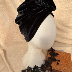 May include: A black velvet turban with a beaded embellishment on the front. The turban is on a white mannequin head.
