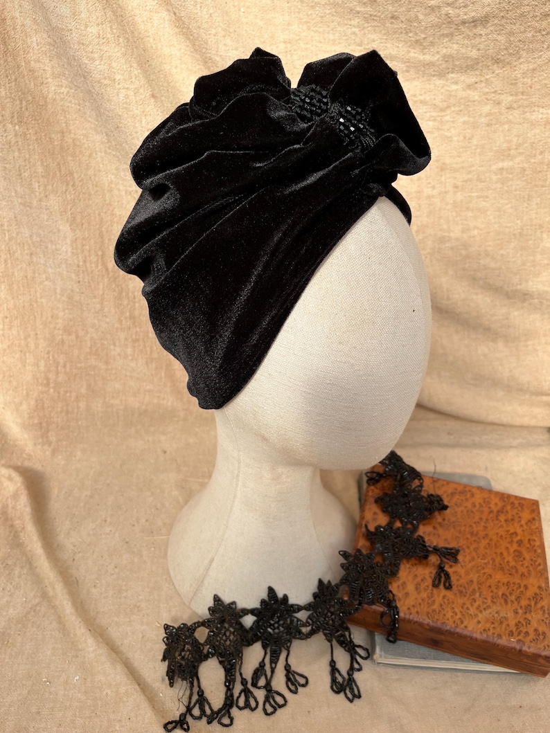 May include: A black velvet turban with a beaded embellishment on the front. The turban is on a white mannequin head.