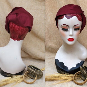 May include: A burgundy satin hat with a bow on the side, displayed on a mannequin head. The hat is on a light brown background with wheat stalks and two woven baskets.