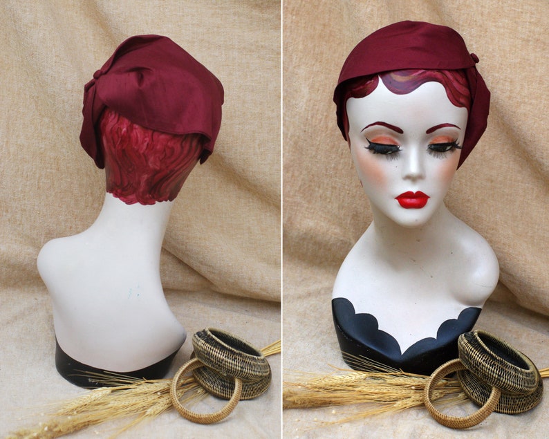 May include: A burgundy satin hat with a bow on the side, displayed on a mannequin head. The hat is on a light brown background with wheat stalks and two woven baskets.