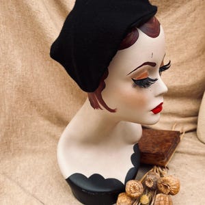 K&ouml;nnte beinhalten: Ein schwarzer Filz-Barett auf einem Mannequin-Kopf. Das Mannequin hat rote Haare und tr&auml;gt Make-up. Der Barett sitzt auf einem schwarz-wei&szlig;en St&auml;nder.