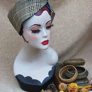 May include: A mannequin head wearing a plaid and teal fabric hat with a brown button. The mannequin is wearing a black and white patterned dress. The hat is sitting on a book with woven bracelets and dried flowers.