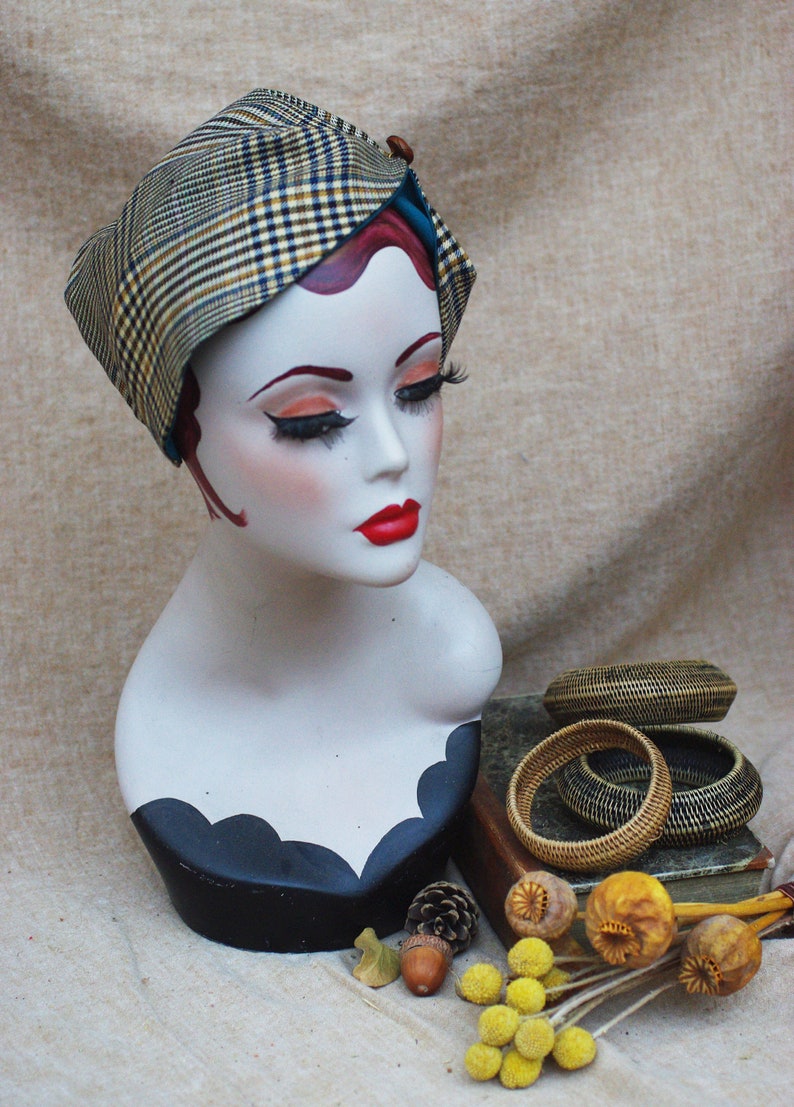 May include: A mannequin head wearing a plaid and teal fabric hat with a brown button. The mannequin is wearing a black and white patterned dress. The hat is sitting on a book with woven bracelets and dried flowers.