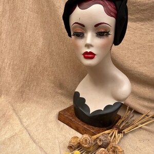 K&ouml;nnte beinhalten: Ein schwarzer Filzhut im Cloche-Stil mit schlichtem Design. Der Hut sitzt auf einem Mannequin-Kopf mit einer schwarz-wei&szlig; bemalten Basis.